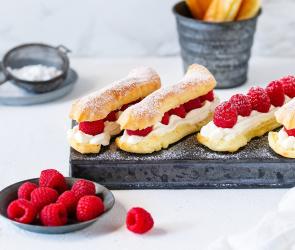 Eclairs auf Servierbrett mit Puderzucker im Sieb und Himbeeren in Schale