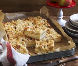 Eine eckige Backform mit Backpapier ausgelegt. Darauf befindet sich ein Birnenkuchen mit Streuseln. Im Vordergrund befinden sich geschnittene Stücke.