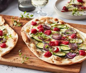 Flammkuchen belegt mit Himbeeren, Zucchini und Ziegenkäse auf einem Holzbrett.