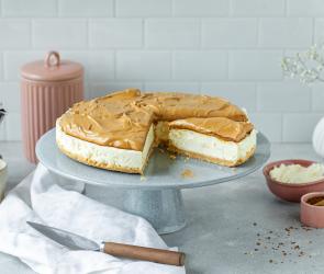 Cheesecake mit Dalgona Topping auf einer Tortenplatte, angeschnitten.