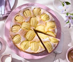 Apfelkuchen auf einem rosa Teller, ein Stück ist angeschnitten und wird herausgenommen.