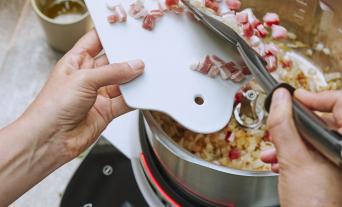 Speck wird zu den gedünsteten Zwiebeln im Cookit Topf zugegeben.