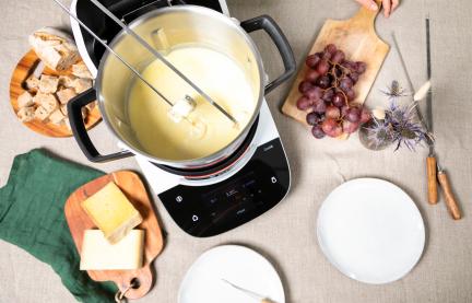 Klassisches Schweizer Käsefondue im Cookit auf einem Tisch, drumherum Zutaten, Käsestücke, Baguette und Trauben