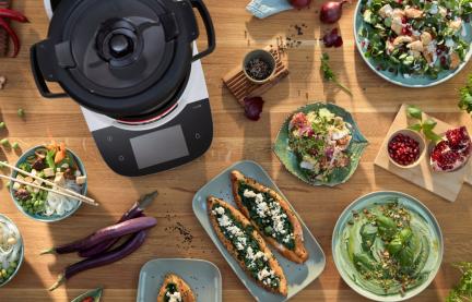 Der Cookit auf einer Tischplatte, daneben verschieden Gerichte, wie Spinat-Pita, Hummus und Couscous-Salat. 