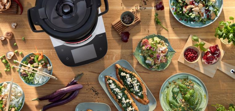 Der Cookit auf einer Tischplatte, daneben verschieden Gerichte, wie Spinat-Pita, Hummus und Couscous-Salat. Der Cookit auf einer Tischplatte, daneben verschieden Gerichte, wie Spinat-Pita, Hummus und Couscous-Salat.