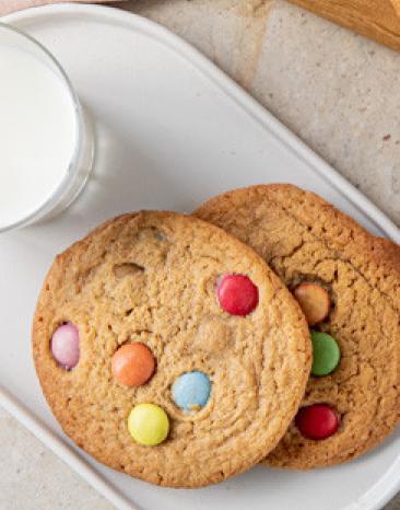 Cookies mit bunten Schokolinsen auf zwei Teller mit einem Glas Milch.