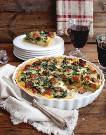 Quiche in der ein Stück fehlt, welches im Hintergrund auf einem Teller liegt.