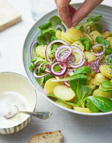 Kartoffelsalat mit Nussbutter in einer Schüssel, eine Hand streut Salz darüber.