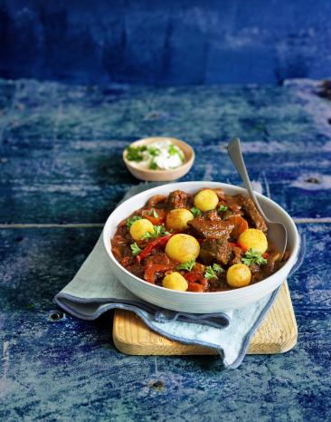 Eine Suppenschüssel mit Gulasch und Polentaklösschen und einem Löffel auf einem Holzbrett.