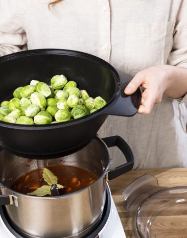 Frau setzt Dampfgaraufsatz gefüllt mit Rosenkohl auf den Cookit Kochtopf