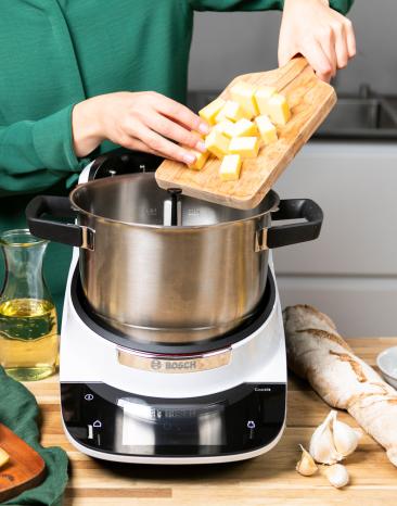 Frau schiebt Käsewürfel von einem Holzbrett in den Cookit Kochtopf