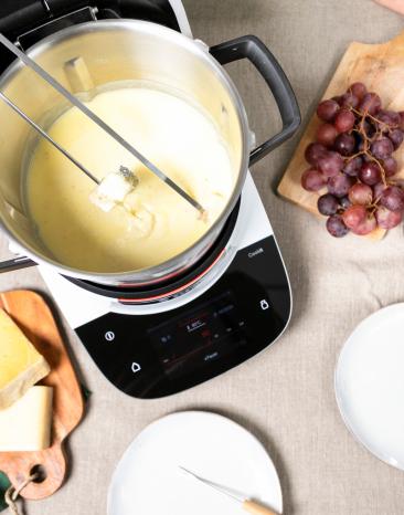 Klassisches Schweizer Käsefondue im Cookit auf einem Tisch, drumherum Zutaten, Käsestücke, Baguette und Trauben