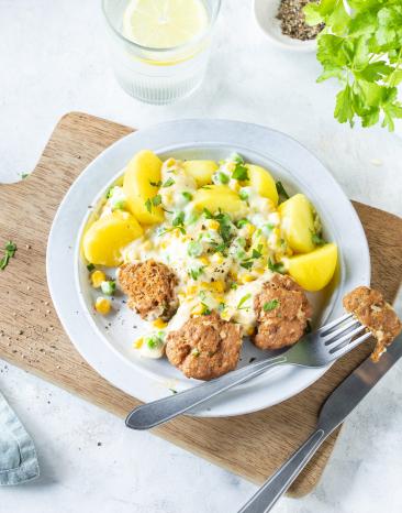 Erbsen und Mais in cremiger Sauce mit Kartoffeln und Frikadellen auf einem Teller, der auf einem Holzbrett steht.