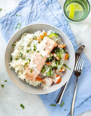 Reis mit Lachsfilet und Gemüse in Rahmsauce in einer Schüssel auf einem blauen Tuch.