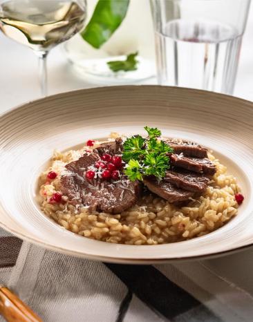 Risotto mit Pilzen und Rindfleischstreifen in einem tiefen Teller, mit Weißweinglas.