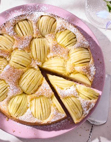 Apfelkuchen auf einem rosa Teller, ein Stück ist angeschnitten und wird herausgenommen.