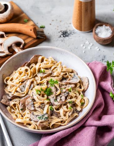 Bandnudeln mit Pilzsauce und Kräutern in einem tiefen Teller, daneben ein Brett mit Pilzen.